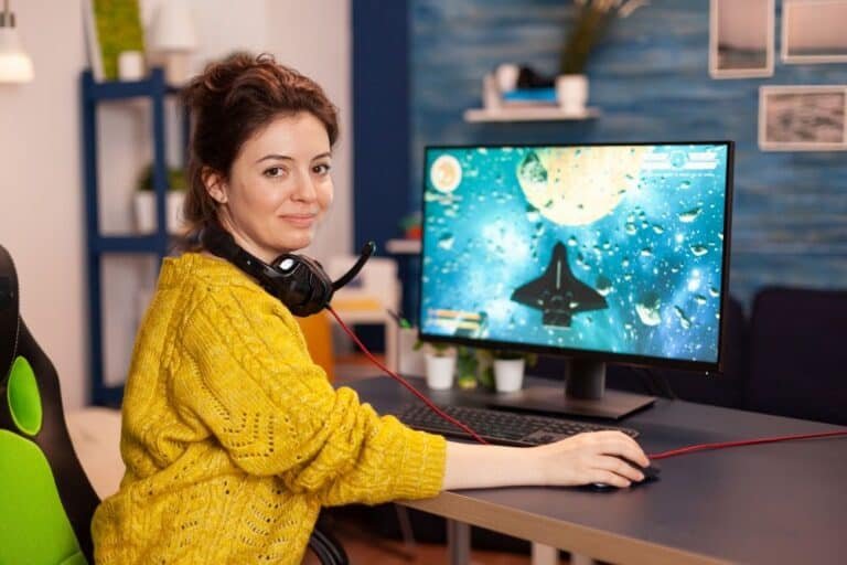 Mujer sentada delante de su ordenador mirando a cámara y con un videojuego en marcha en su monitor.